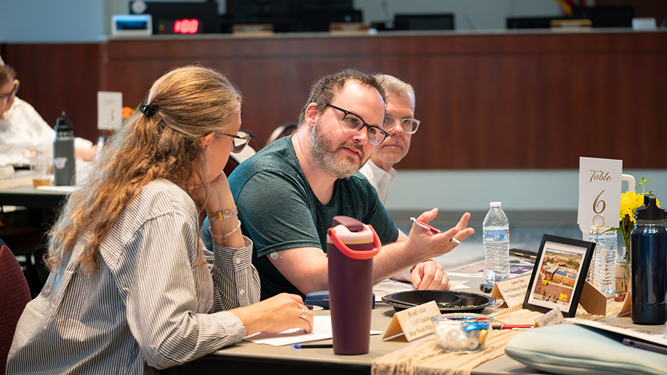 People seated around a conference table during a meeting. In the foreground, one participant gestures while speaking to another participant who is taking notes. Water bottles, notebooks, name tents, and a table marker labeled “Table 6” are visible on the table. A wood-paneled meeting room and additional attendees appear in the background.