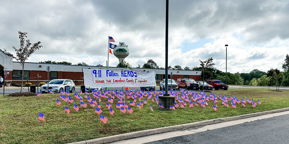 Memorial display with numerous small American flags on grass honoring 9/11 fallen heroes, with a banner thanking Loudoun County responders.