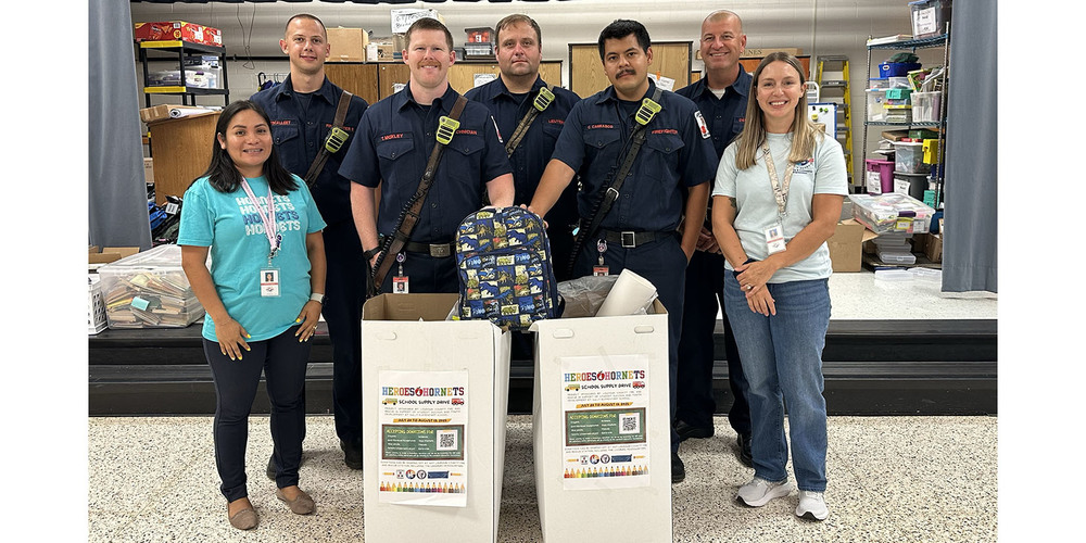 Group of firefighters and two women standing behind donation boxes filled with school supplies in a community center.