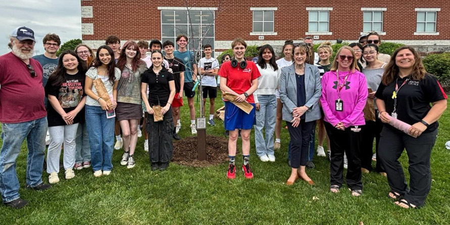 Group of diverse people standing outdoors on grass around a newly planted young tree in front of a brick building under a cloudy sky.