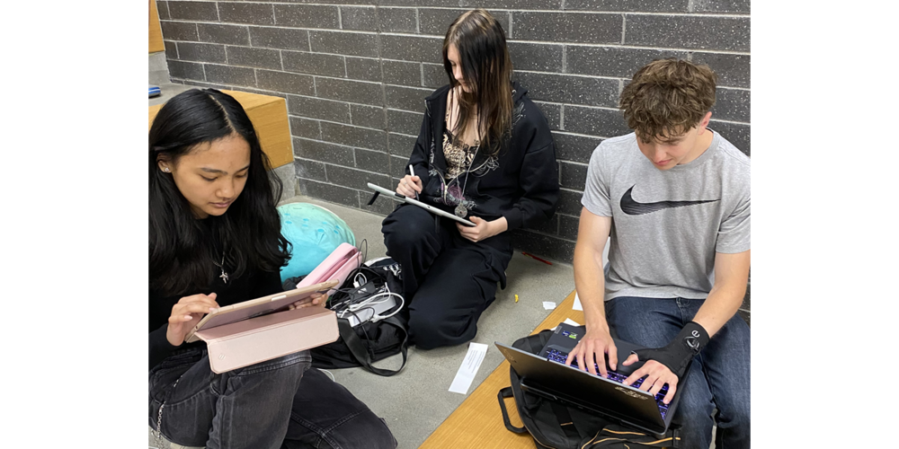 Three people sit on a carpeted floor against a gray brick wall, each using a digital device. One person on the left holds a tablet in a pink case, another in the center writes on a tablet with a stylus, and a third on the right types on a laptop resting on a low wooden surface. Bags, cables, and small personal items are scattered nearby.