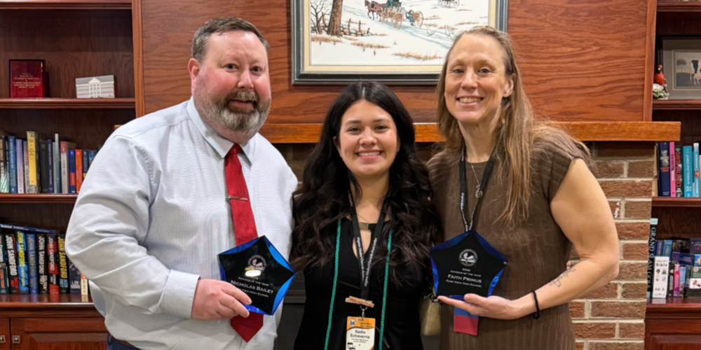 Three people stand indoors in front of bookshelves, each holding a glass-style award. A framed winter landscape artwork hangs on the wall behind them.