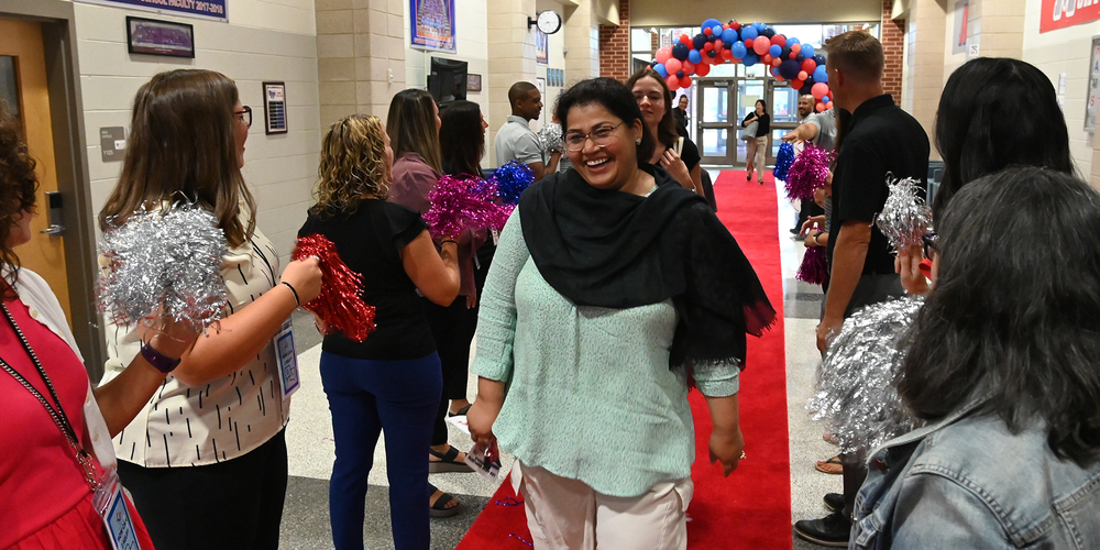 New staff walking down a school hallway on a red carpet surrounded by current staff cheering.
