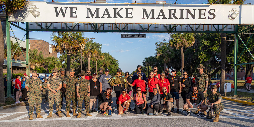Group of US Marines posing with others in the street under a sign reading, "We Make Marines - Clearance 15' 0''."
