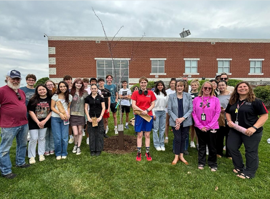 Group of diverse people standing outdoors on grass around a newly planted young tree in front of a brick building under a cloudy sky.