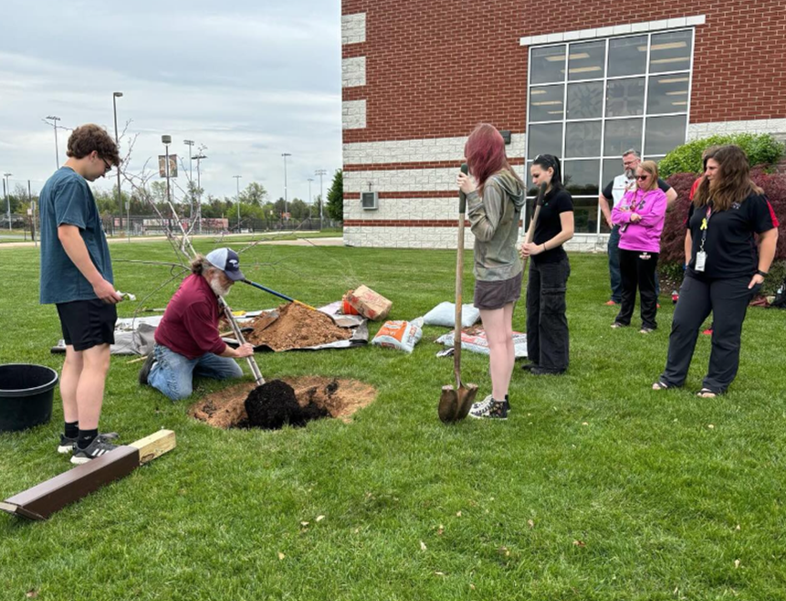 Group of people gathered on a lawn near a brick building, preparing to plant a tree in a freshly dug hole with soil and gardening tools nearby.
