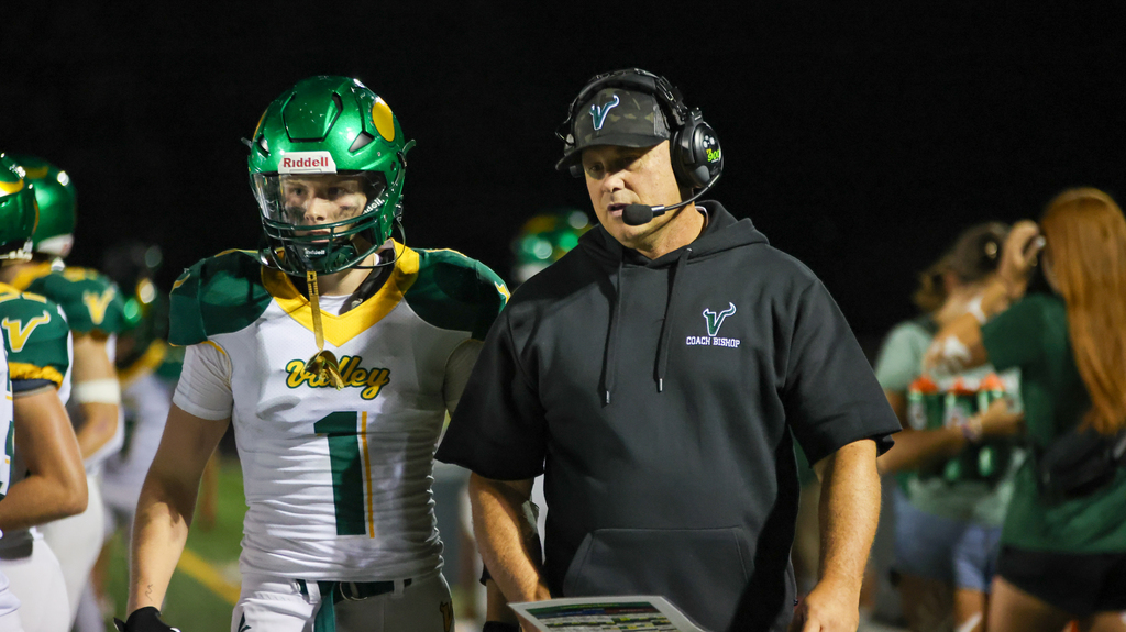  Football player wearing a green helmet and white jersey number 1 stands next to a coach in black hoodie labeled Coach Bishop during a night game.