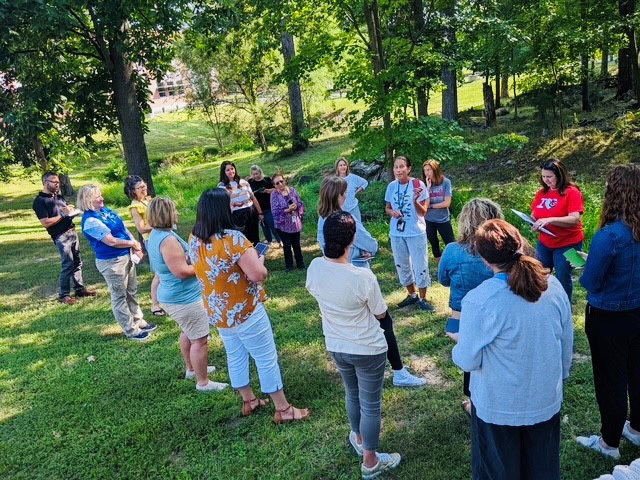 Group of adults standing in a circle outdoors on a sunny day, engaged in conversation in a green park area with trees.