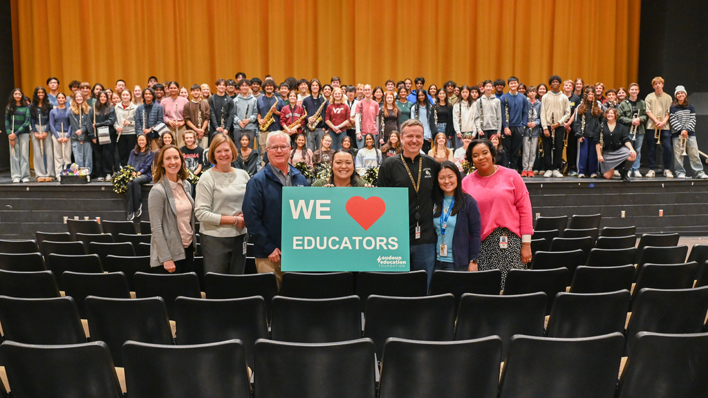 A diverse group of educators and students gathered on a stage, with five adults holding a sign that says 'We ❤️ Educators' in front of empty auditorium seats.
