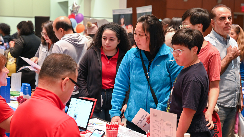 People gathered at a busy indoor event, engaging with representatives at a table with informational materials and a laptop.