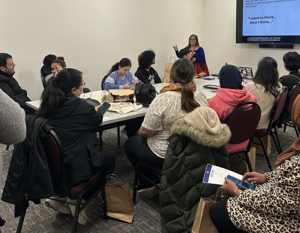 A group of diverse adults attentively listening to a woman presenting in a classroom setting with a slide on the screen.