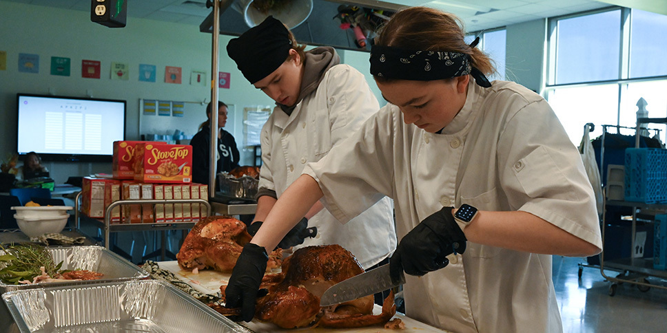 Two culinary students in white chef coats carving roasted turkeys on a cutting board in a classroom kitchen.