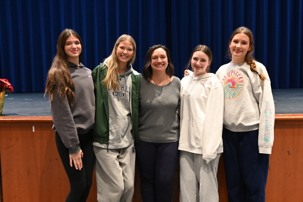 Five casually dressed individuals standing together in front of a stage with a blue curtain backdrop indoors.