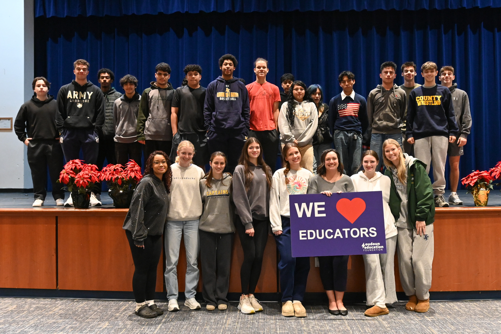 Group of diverse students and educators posing on a stage with a sign reading 'We ❤️ Educators' surrounded by poinsettia plants.