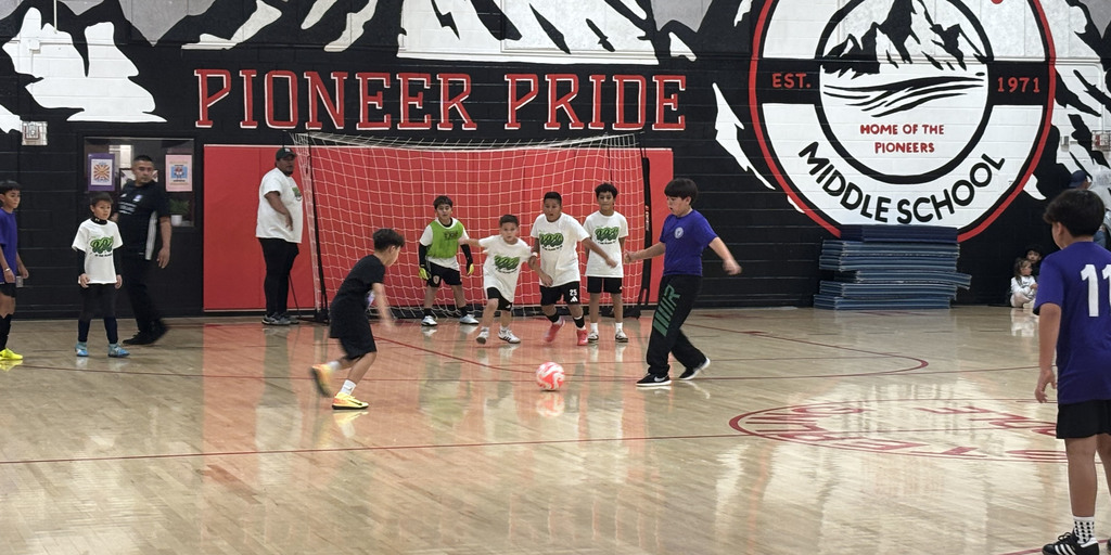 Young boys playing indoor soccer in a middle school gym with a Pioneer Pride banner and mascot logo on the wall.