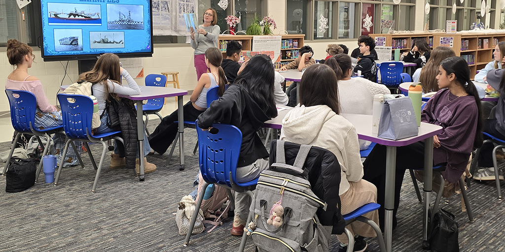 Students seated in a library classroom attentively watching a teacher present ship images on a large screen.