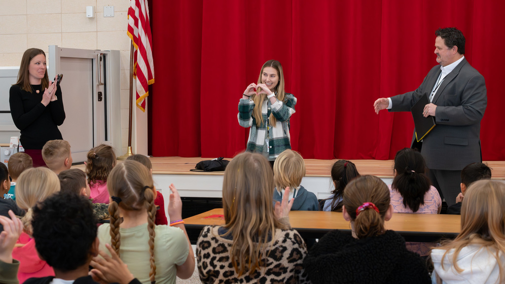 Teacher using sign language to communicate with a group of attentive elementary students in a classroom with a red curtain backdrop.