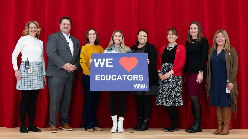 Group of eight people standing in front of a red curtain, holding a sign that says 'We ❤️ Educators'.