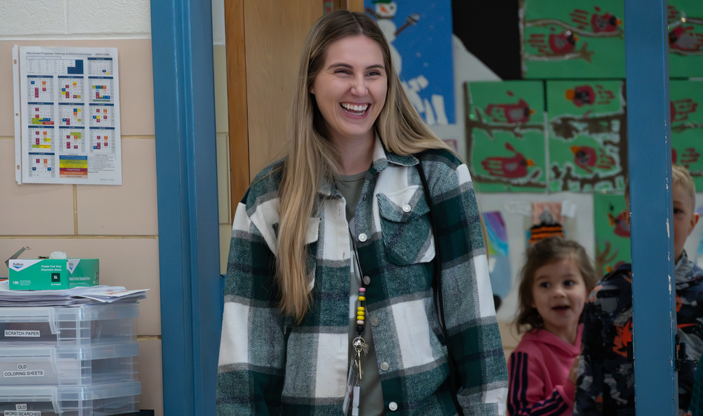Teacher wearing a green and white plaid jacket standing in a classroom with colorful children's artwork on the walls.