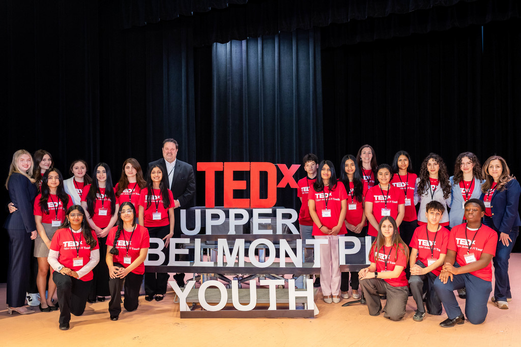 Group photo of diverse youth and adults wearing red TEDx Upper Belmont Place Youth shirts on stage with TEDx sign.
