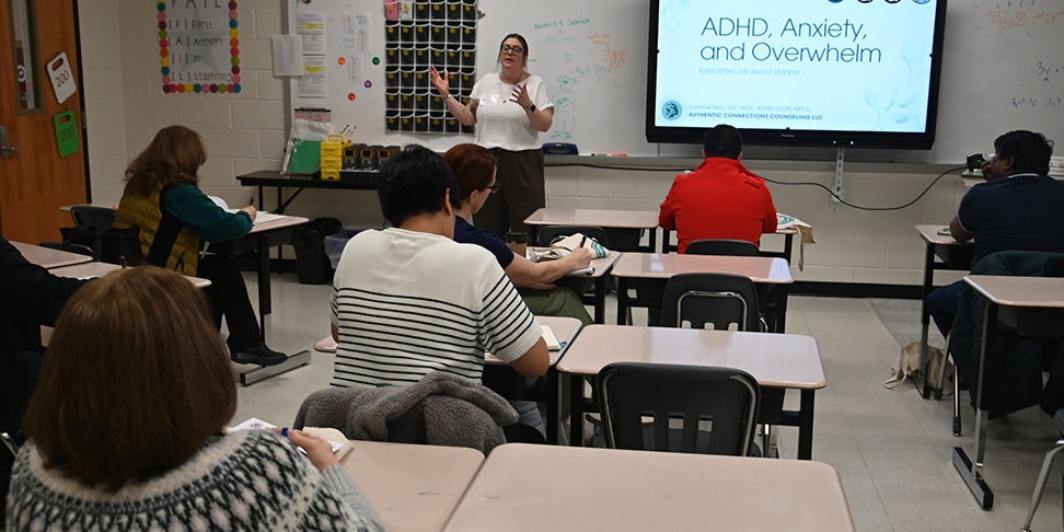 A classroom setting where an instructor stands at the front, giving a presentation titled “ADHD, Anxiety, and Overwhelm” on a large screen. Several attendees sit at desks, taking notes while facing the presenter.