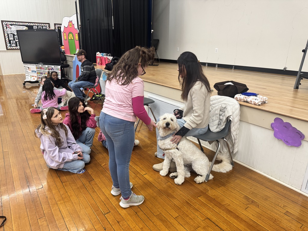 Children sitting on a wooden floor watching a woman seated on a chair petting a white dog near a stage in a gym.