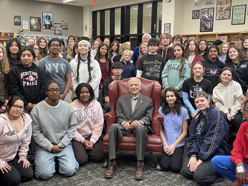 A Holocaust survivor sits in a red armchair surrounded by a large group of students posing for a group photo inside a school library.