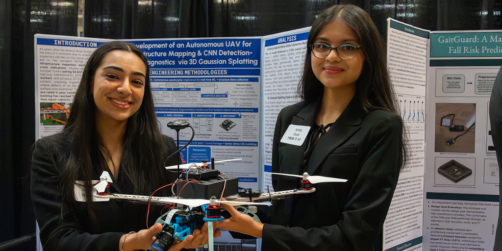 Two female students stand at a research poster session holding a quadcopter drone with a mounted camera, sensors, wiring, and propellers. Behind them are large academic posters with diagrams and text about autonomous UAV development, infrastructure mapping, and machine learning analysis. The scene appears to be an indoor conference or exhibition hall with display boards and technical visuals.