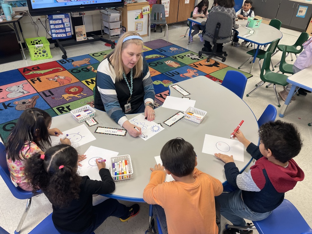 An adult seated at a round classroom table works with a small group of children drawing on paper with markers. Name labels, marker containers, and worksheets are on the table. The classroom includes colorful alphabet rugs, small chairs, and additional groups working in the background.