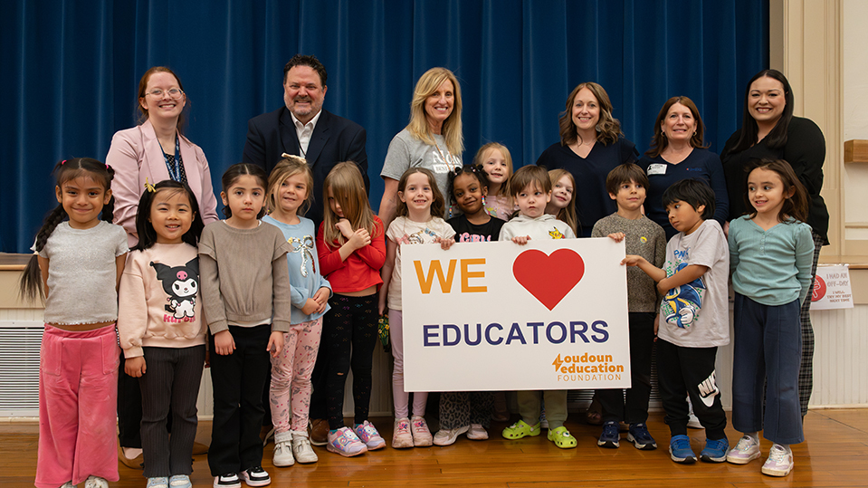 Group of young children standing with several adults on a stage, holding a sign that reads ‘We ♥ Educators’ with the Loudoun Education Foundation logo. Blue stage curtains form the background.