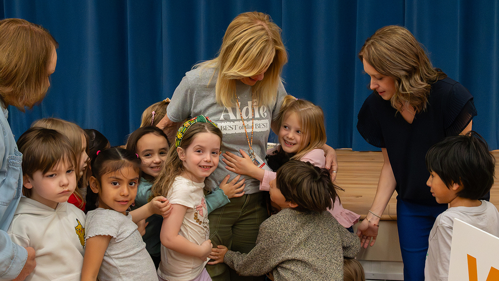 Several young children gathered closely around an adult standing on a stage, with other adults nearby. The setting includes a wooden stage floor and blue curtains in the background.