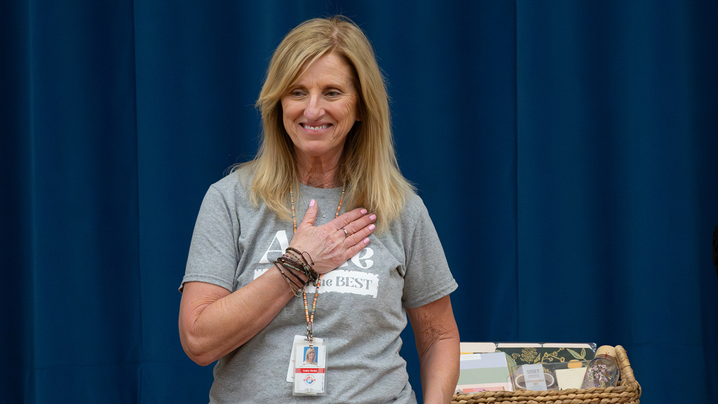 An adult standing on a stage in front of blue curtains, wearing a gray T‑shirt with the word ‘Aldie’ visible and a lanyard badge, with a basket of items on a table beside the person.