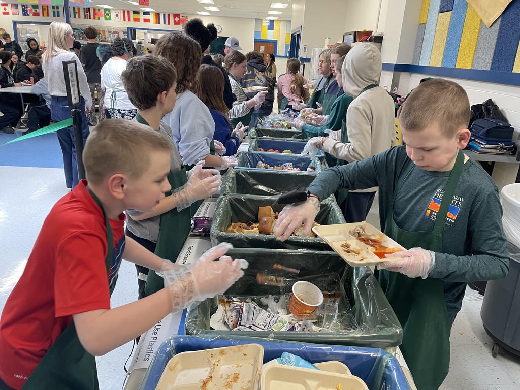 Students wearing aprons and disposable gloves stand along a cafeteria sorting station, placing leftover food and containers into large labeled bins. Other students wait in line behind the station, and international flags hang from the ceiling in the background.