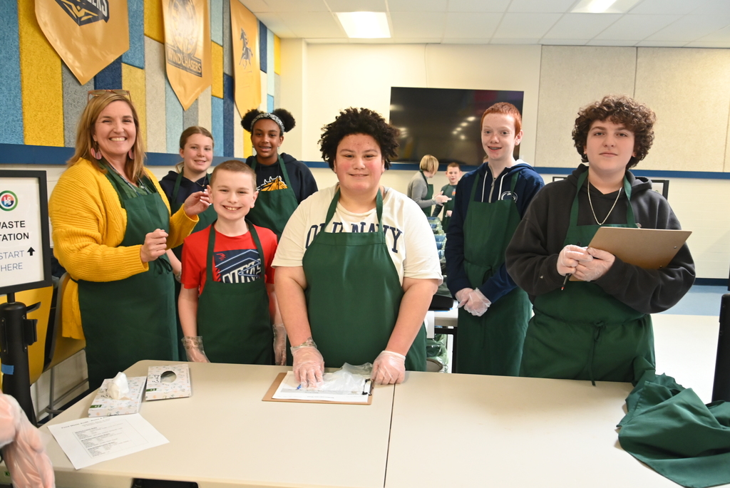 A group of students and one adult stand behind a table in a school cafeteria, wearing green aprons and gloves. Clipboards and papers are on the table, and a sign in the background reads “Waste Station,” with cafeteria tables and wall banners visible behind them.