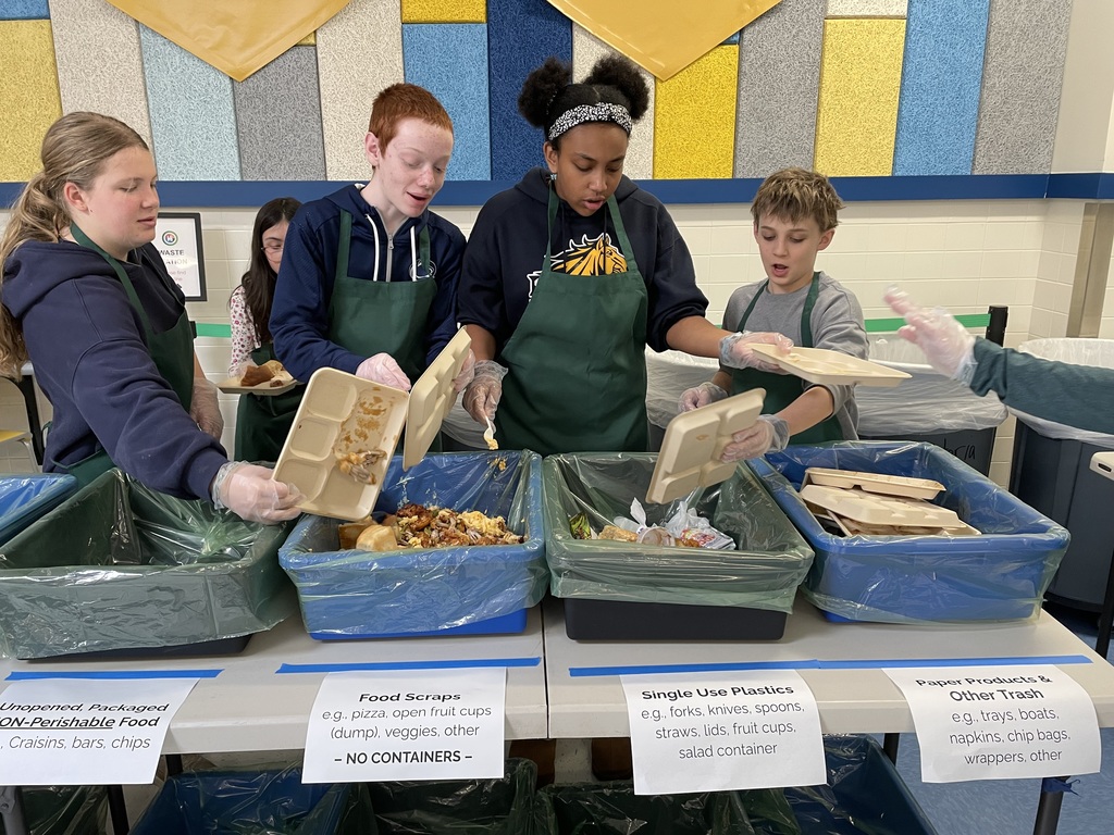 Several students wearing aprons and gloves sort cafeteria waste into color-coded bins labeled “Food Scraps,” “Single Use Plastics,” and “Other Trash,” placing leftovers from divided trays into the appropriate containers. Informational signs are taped to the tables above each bin.
