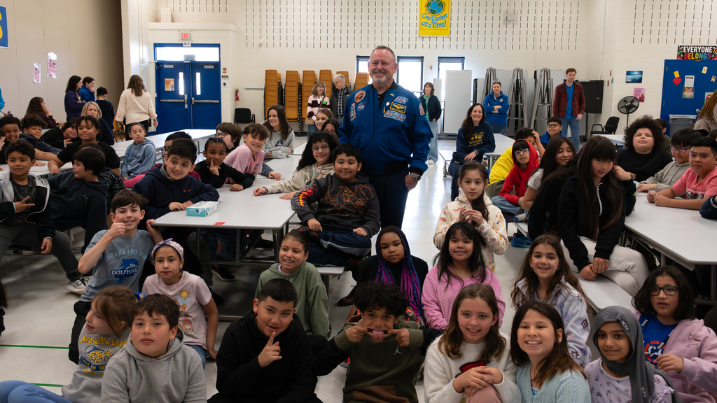 A person wearing a blue astronaut-style flight suit stands among rows of seated elementary-age students gathered around cafeteria tables in a school gym or multipurpose room, with teachers and school staff visible in the background.