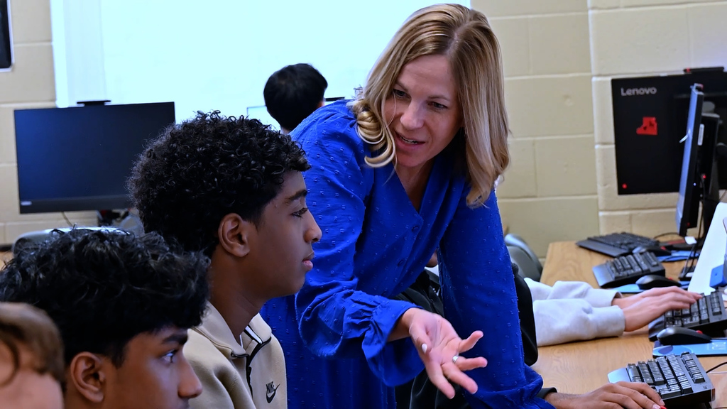 An adult leaning over a row of seated students in a computer lab, gesturing toward a desktop monitor while the students work at keyboards. Multiple desktop computers are visible on long tables, and several faces are intentionally blurred.