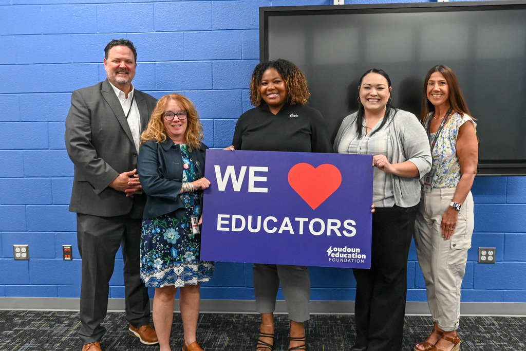Five people stand in a row indoors against a blue cinderblock wall and a large black display screen. Two people in the center hold a large purple sign that reads “WE ♥ EDUCATORS” with “Loudoun Education Foundation” printed in the lower corner. The group is dressed in business and casual attire, and all wear ID badges or lanyards.