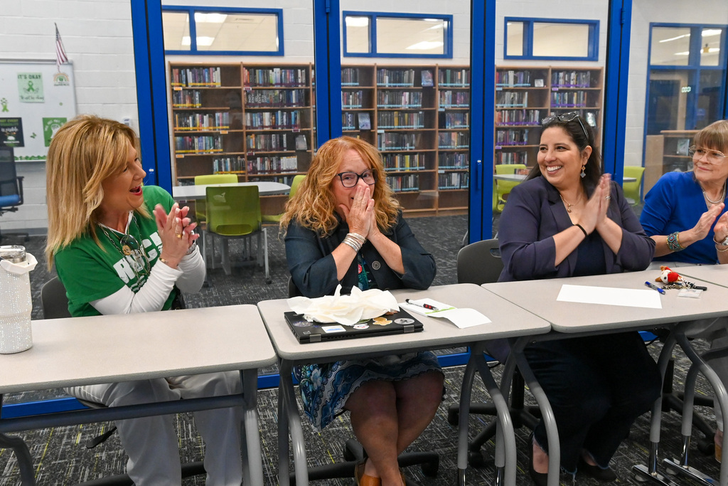 Four people sit at desks in a classroom or library space with glass windows revealing bookshelves behind them. The person in the center, with curly hair and glasses, holds both hands near the face while others on either side clap.