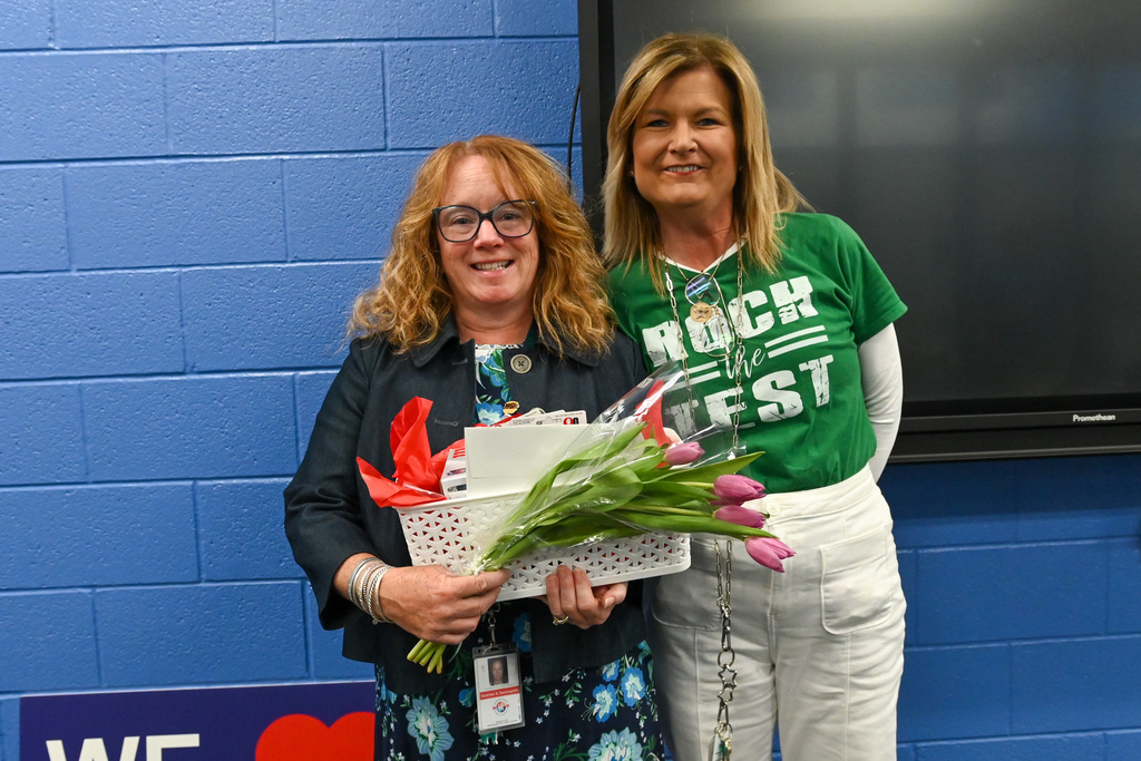 Two people stand side by side indoors against a blue cinderblock wall and a black display screen. The person on the left holds a white basket filled with items and a bouquet of pink tulips. Both people wear layered outfits, and ID badges hang