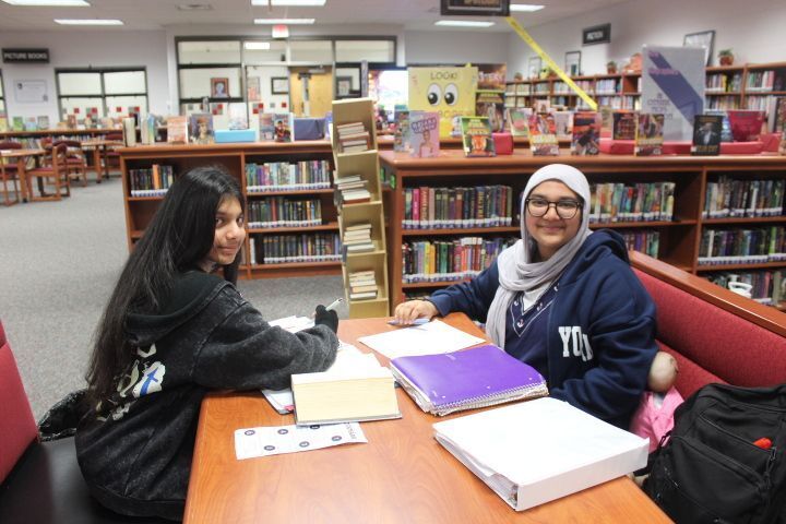 Two students study together at a library table surrounded by bookshelves filled with books.