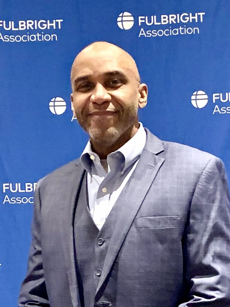 Dr. Marcus White in a gray suit and white shirt smiling in front of a blue Fulbright Association backdrop.