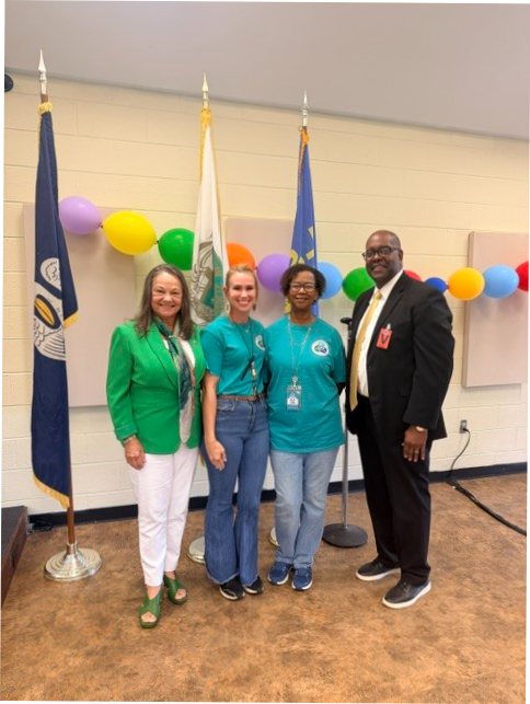 Sandy Hollaway, Bernel Cook, Alisha Butler and Dr. Shirley Lewis,  pose for a photo at a back-to-school event. Two individuals wear teal event T-shirts, one wears a green blazer with white pants, and one wears a dark suit with a light-colored tie. The group stands between flag displays, with a colorful balloon garland decorating the background. The photo was taken inside a meeting space with neutral-colored walls and flooring.