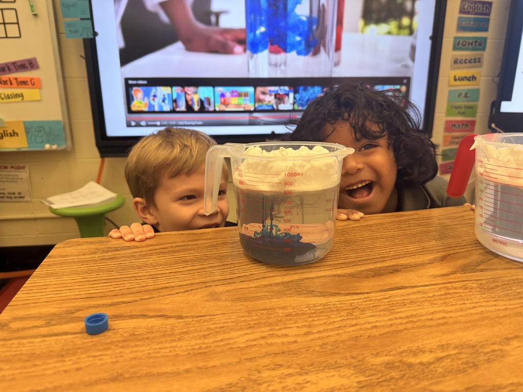 Two students smiling looking at a measuring cup with water, food coloring, and shaving cream