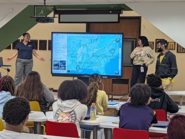Three people standing next to a promethean board for a presentation.