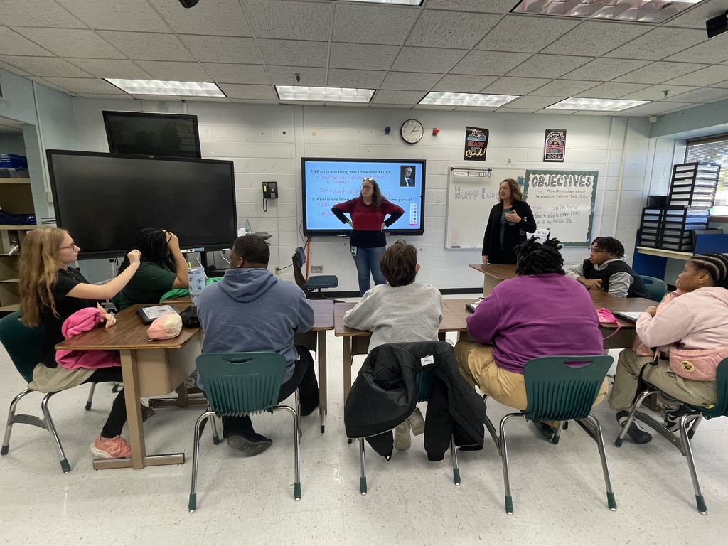 Teacher standing at the board, students sitting at desks looking at the board