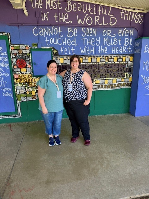 Two staff standing and smiling in front of a colorful painted wall. 