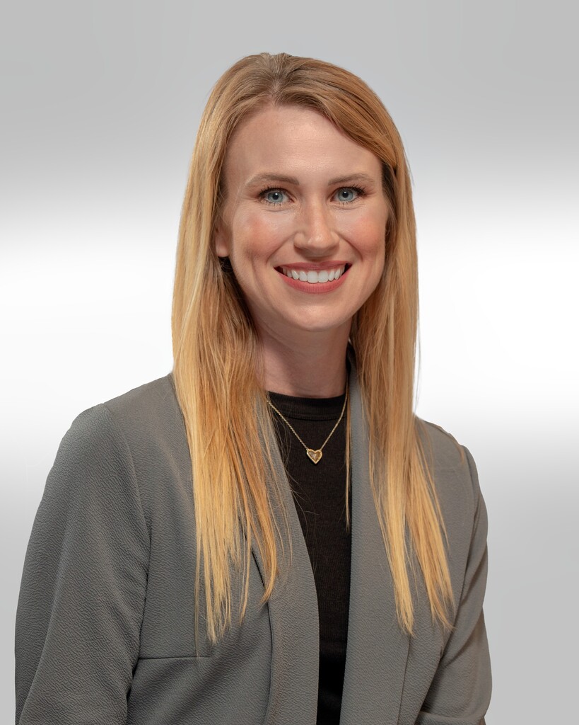 Studio-style portrait of Ms. Alisha Butler with long light-colored hair, wearing a gray blazer over a dark shirt and a heart-shaped pendant necklace. She is centered in the image, facing the camera, with a plain light gray background.