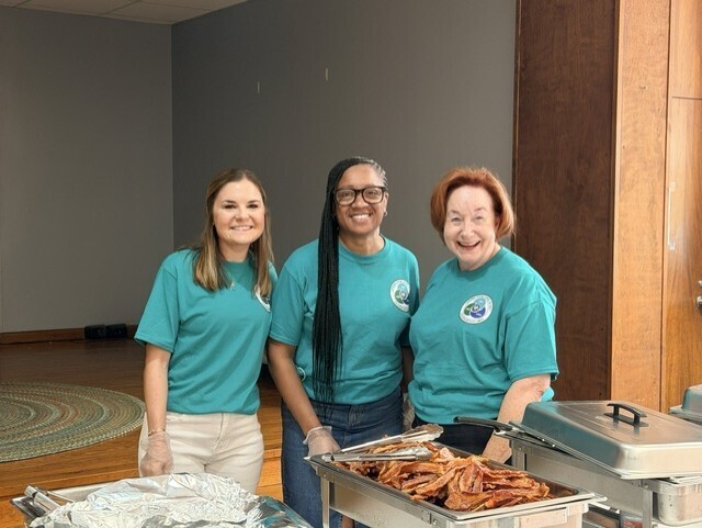 Diana Goff, Perpetra Frazier and Kat Carr , wearing matching teal "Students First" T-shirts stand behind a breakfast serving station at a back-to-school event. In front of them are chafing dishes and trays of breakfast food, including bacon. The group poses for a photo inside a meeting room with wood-paneled walls and natural light coming from the right side of the room.