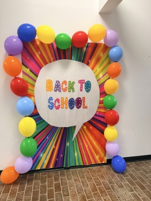 A colorful back-to-school photo backdrop stands against a wall inside an event venue. The display features a rainbow-striped background with a white speech bubble in the center containing the words “BACK TO SCHOOL” in multicolored letters. Bright balloons in purple, blue, green, yellow, orange, and red frame the display on all sides, creating a festive welcome area for attendees.